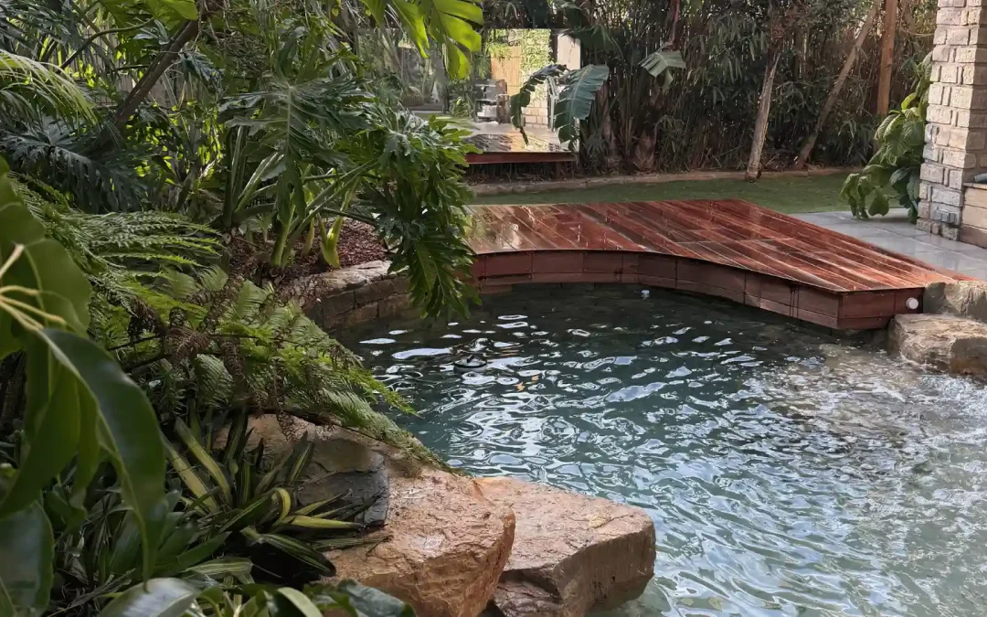 Création d'une piscine lagon naturelle dans le Var, avec plage de sable immergée, gros rochers décoratifs et ponton en bois courbé, entourée d'une végétation tropicale dense.
