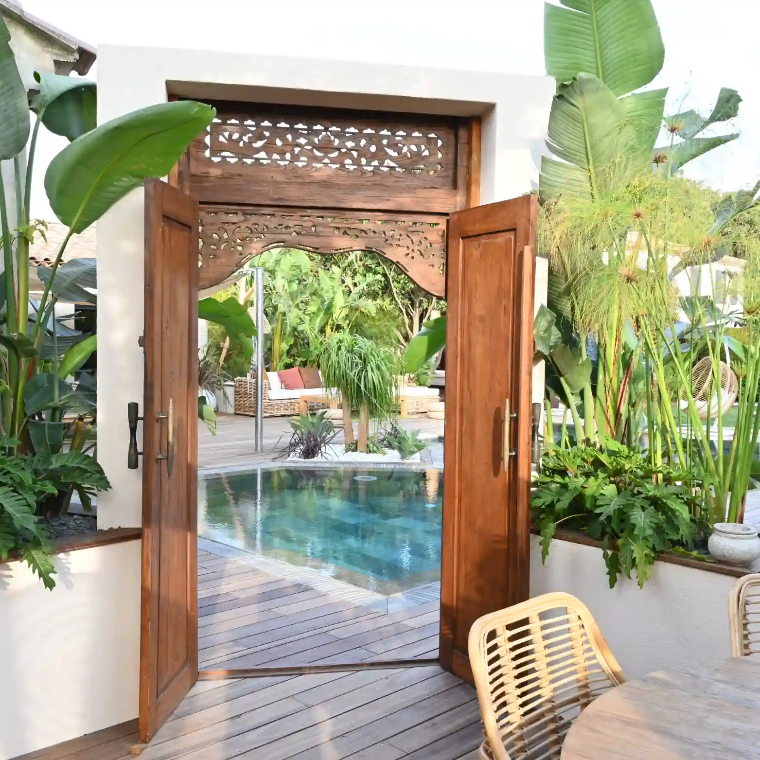 Création paysagère d'exception à Hyères : porte balinaise en bois sculpté ouvrant sur une piscine carrelée et une terrasse en bois, entourée de bananiers et papyrus géants.