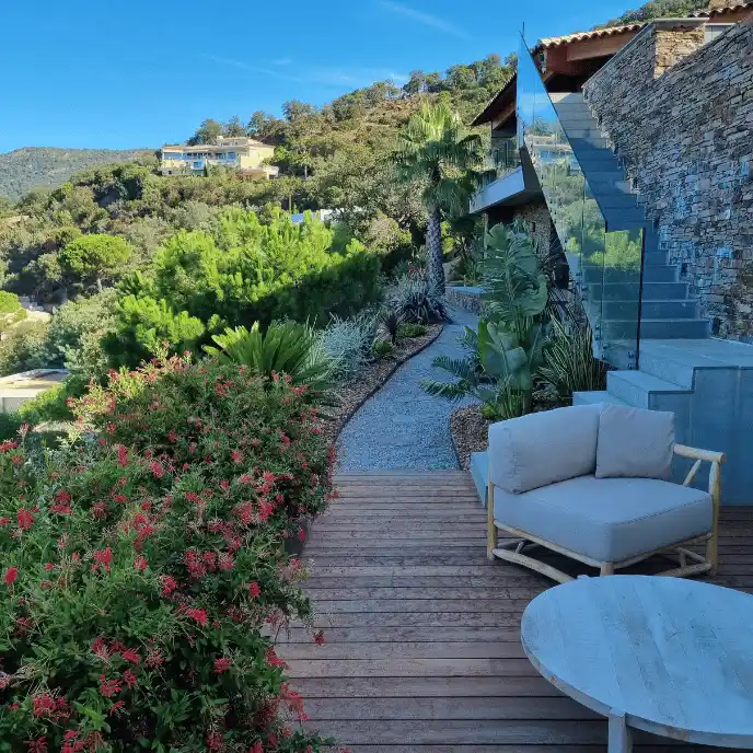 Création d'un jardin exotique dense sur un terrain en pente : cheminement en gravier traversant une végétation luxuriante (Palmiers, Strelitzias, arbustes rouges) longeant une terrasse en bois moderne.