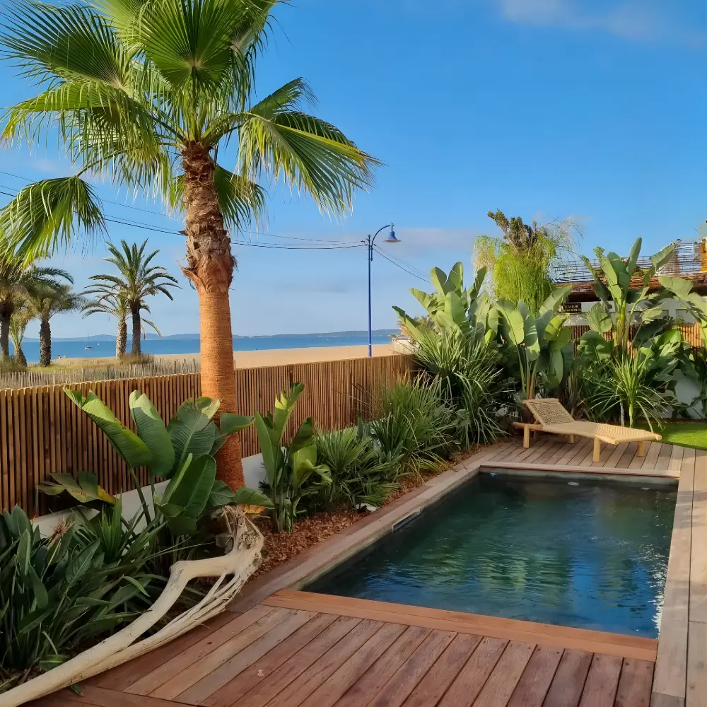 Aménagement paysager en bord de mer à Hyères : petite piscine rectangulaire entourée d'une terrasse en bois et de palmiers, avec vue directe sur la plage.