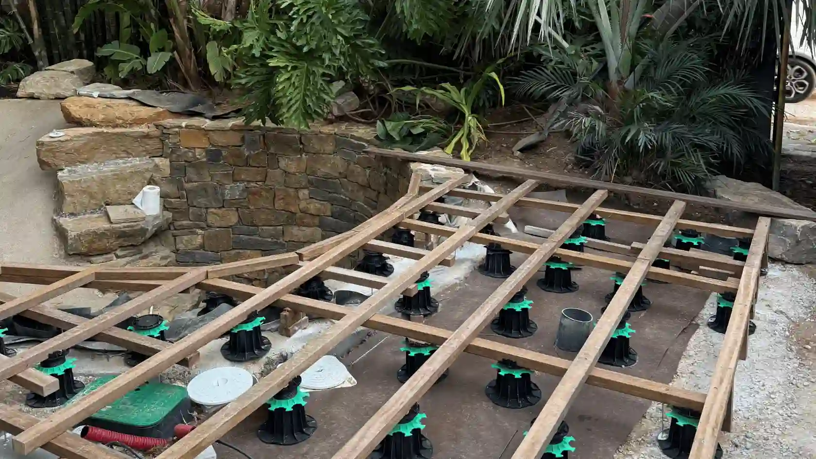 Construction de la structure d'une terrasse en bois exotique sur plots réglables et lambourdes, bordant un mur en pierre dans un aménagement de lagon tropical.