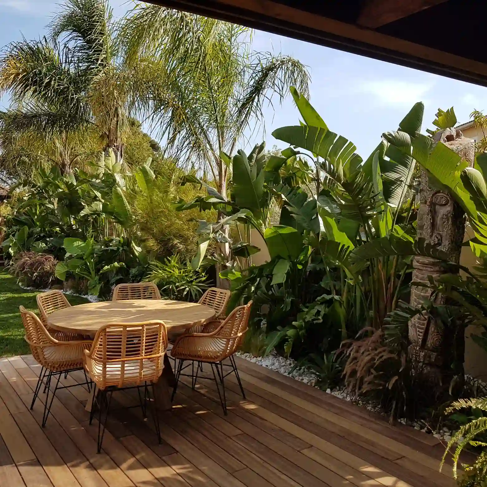 Aménagement de terrasse en bois à Hyères : coin repas convivial avec table ronde et chaises en rotin, niché au cœur d'un jardin exotique luxuriant avec statue totem.