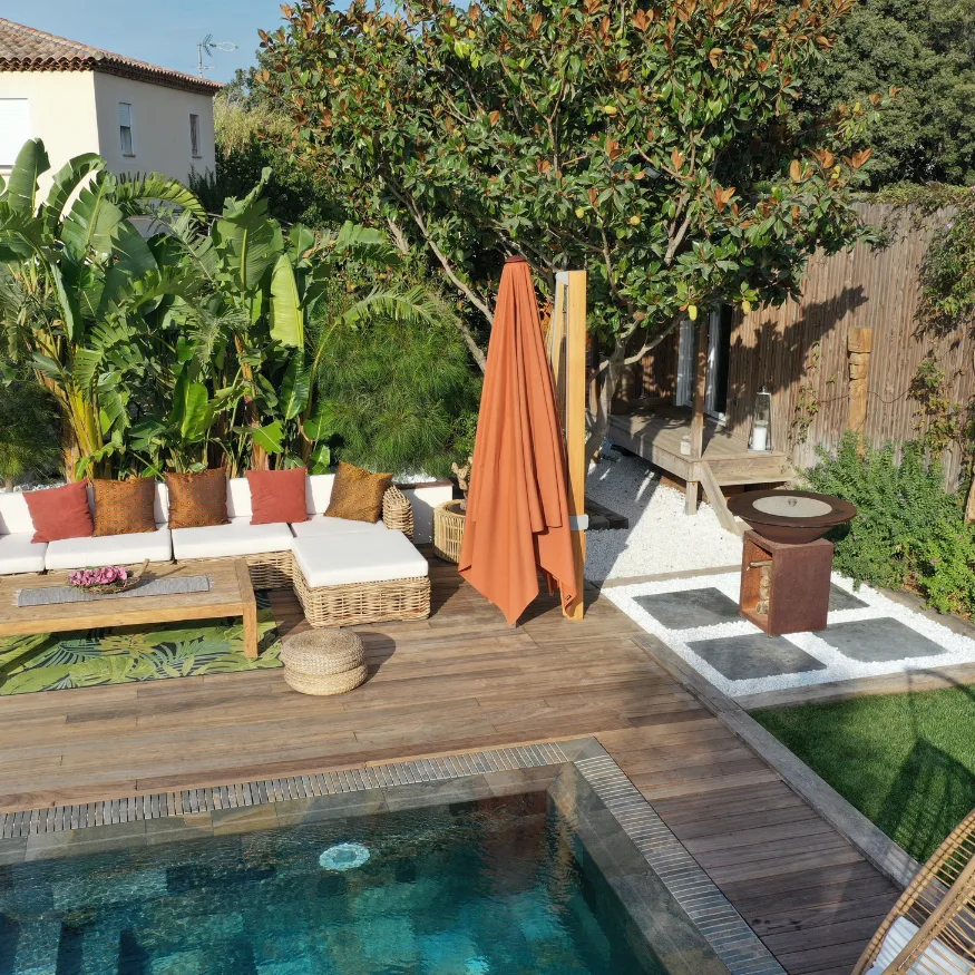 Aménagement extérieur complet à Hyères : terrasse en bois avec salon de jardin cosy, piscine carrelée, brasero en acier Corten sur graviers blancs et bananiers.