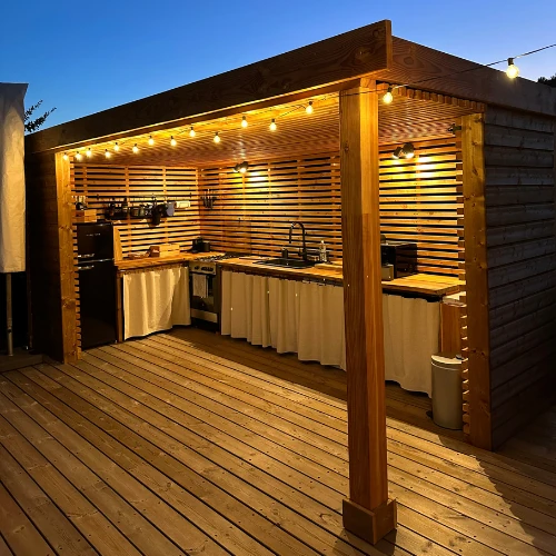 Conception d'une cuisine d'été sur mesure sous pergola en bois avec éclairage guirlande guinguette. Aménagement extérieur complet avec terrasse bois et rangements intégrés.