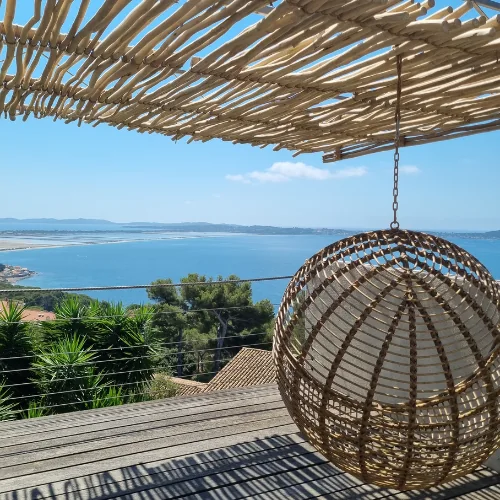 Terrasse en bois avec pergola en bois naturel offrant une vue panoramique sur la mer à Hyères. Aménagement cocooning avec fauteuil suspendu en rotin sous un ombrage ajouré.